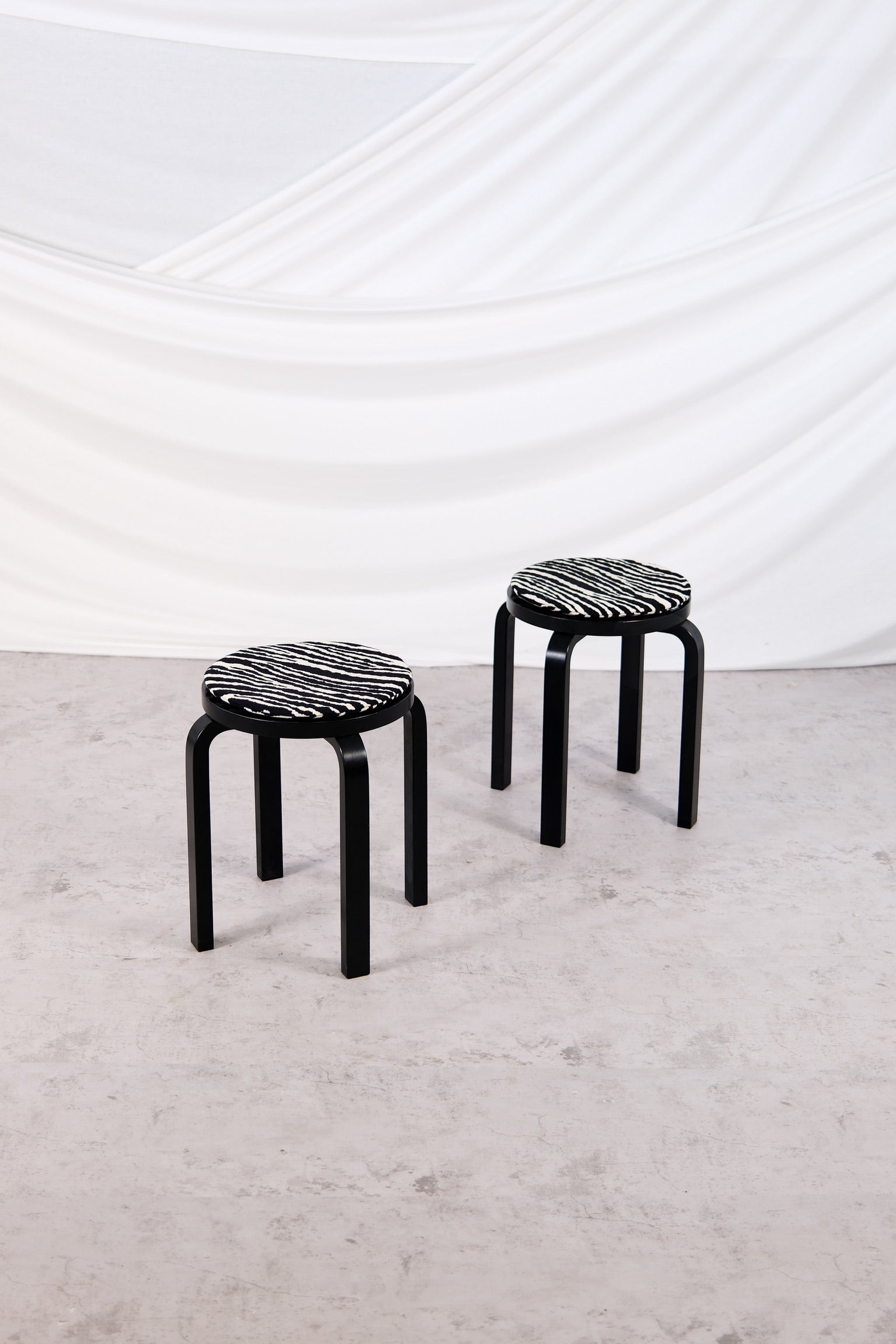 "Pair of black Artek E60 stools with zebra-patterned cushions, design by Alvar Aalto, photographed in studio setting."