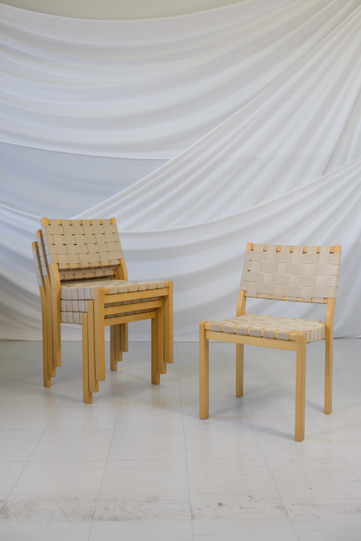 Set of four vintage Artek 611 chairs designed by Alvar Aalto with birch frames and linen webbing, visible wear and staining, photographed in front of a white fabric backdrop.
