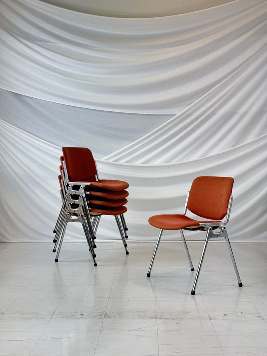 Stackable DOC 106 chairs by Giancarlo Piretti for Castelli, upholstered in burnt orange fabric with chrome legs, shown in a group of six with one separate chair in front of a white draped backdrop.