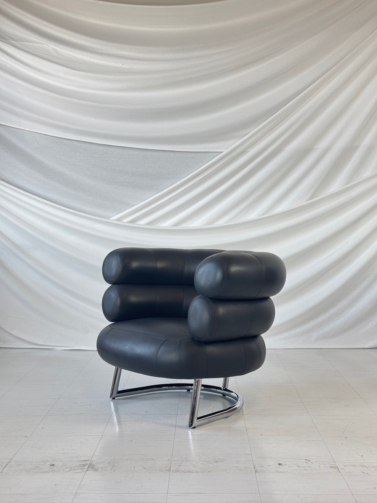Eileen Gray Bibendum chair in black leather with chrome base, sculptural tubular design, photographed against draped fabric backdrop