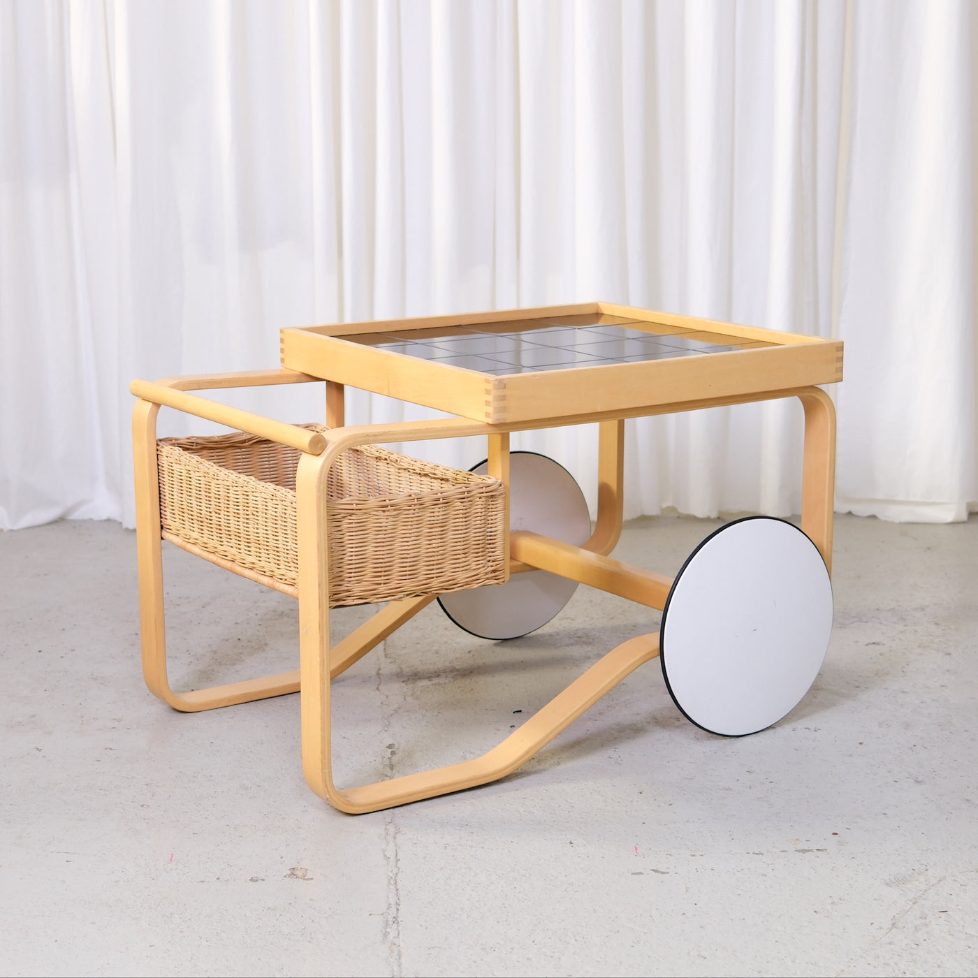 Artek 900 tea trolley by Alvar Aalto with black ceramic tiles, bent birch frame, woven basket and white wheels.