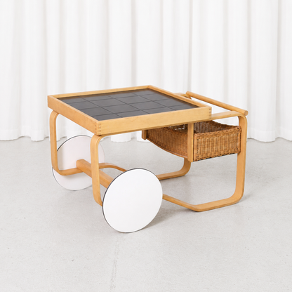 Vintage Artek 900 Tea Trolley designed by Alvar Aalto in 1937, featuring a bent birch frame, black ceramic tiles, rattan basket and white wheels.
