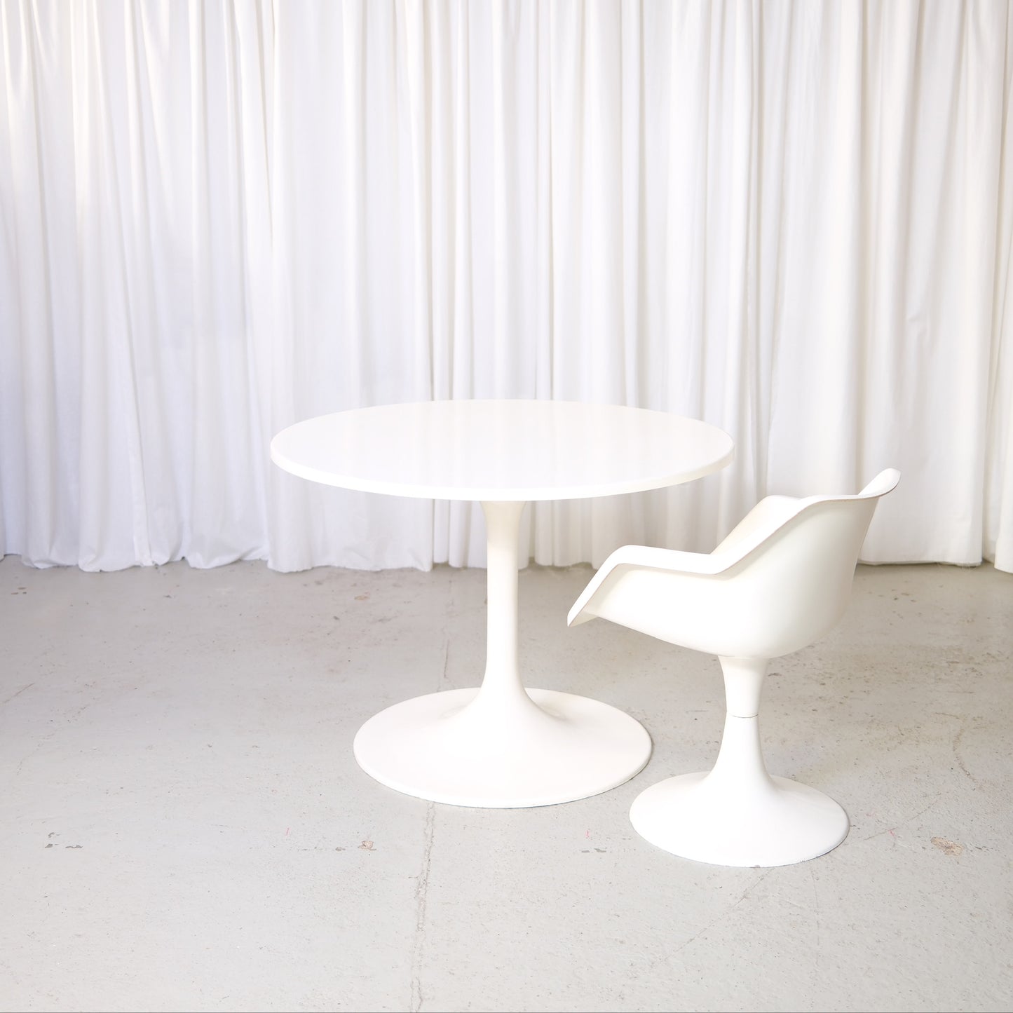 Vintage Asko Amigo round white dining table with pedestal base in front of white curtains.