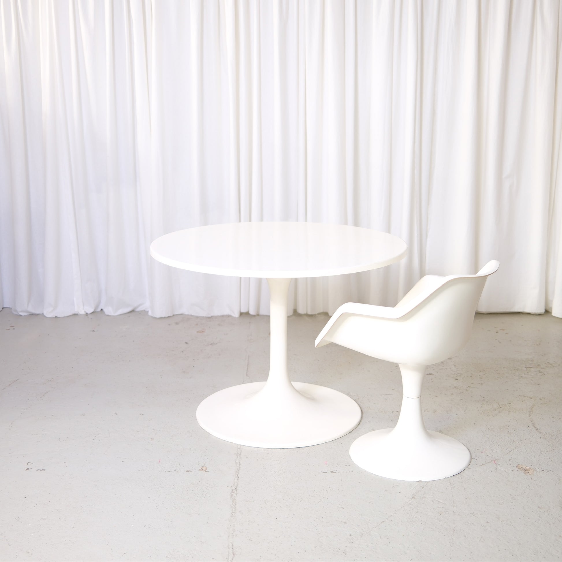 Vintage Asko Amigo round white dining table with pedestal base in front of white curtains.