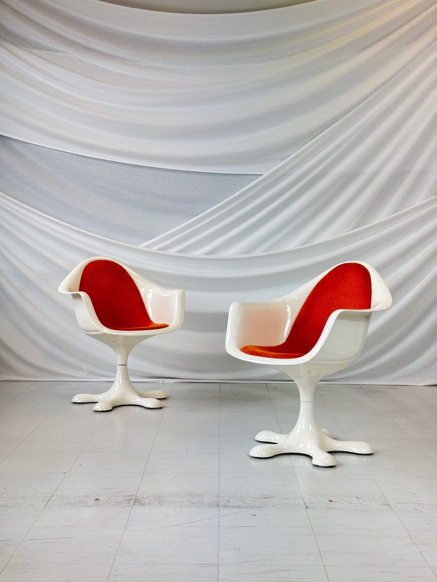 Set of five vintage Orion chairs by Eero Aarnio with white fiberglass frames and bright orange upholstery, featuring star-shaped swivel bases, arranged in front of a white draped backdrop.