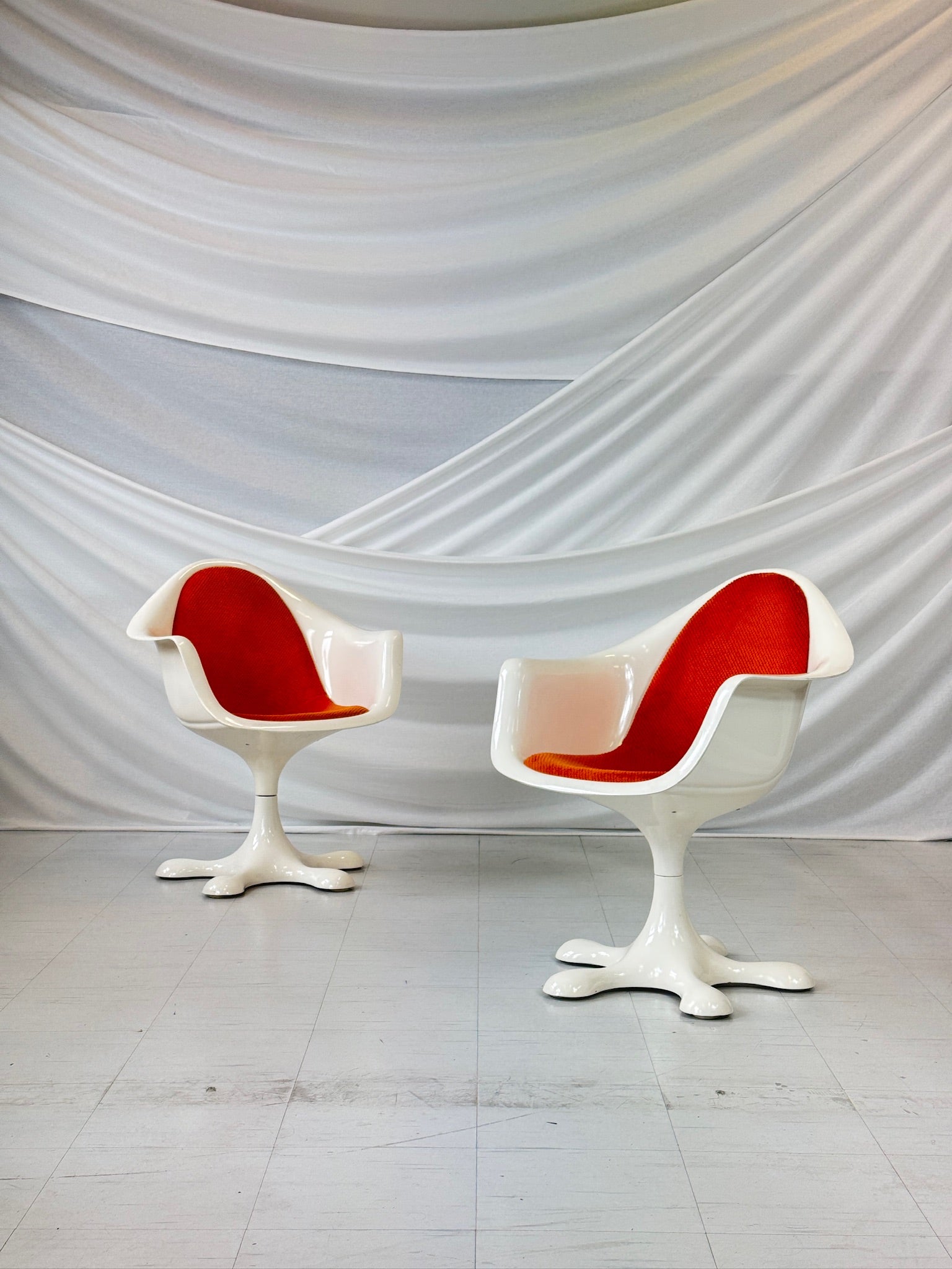 Set of five vintage Orion chairs by Eero Aarnio with white fiberglass frames and bright orange upholstery, featuring star-shaped swivel bases, arranged in front of a white draped backdrop.