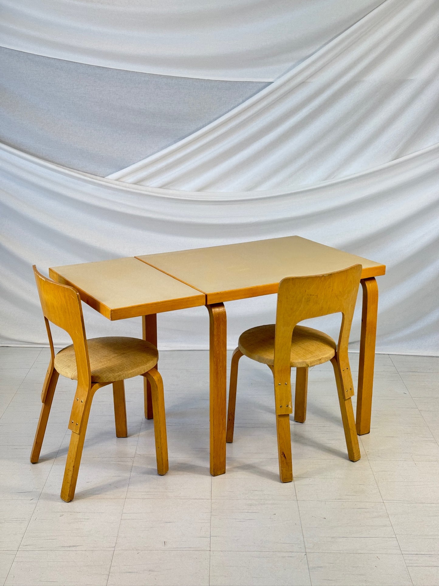 Vintage Artek DL82 table designed by Alvar Aalto, with birch frame and one drop-leaf side extended, photographed against a white draped backdrop.
