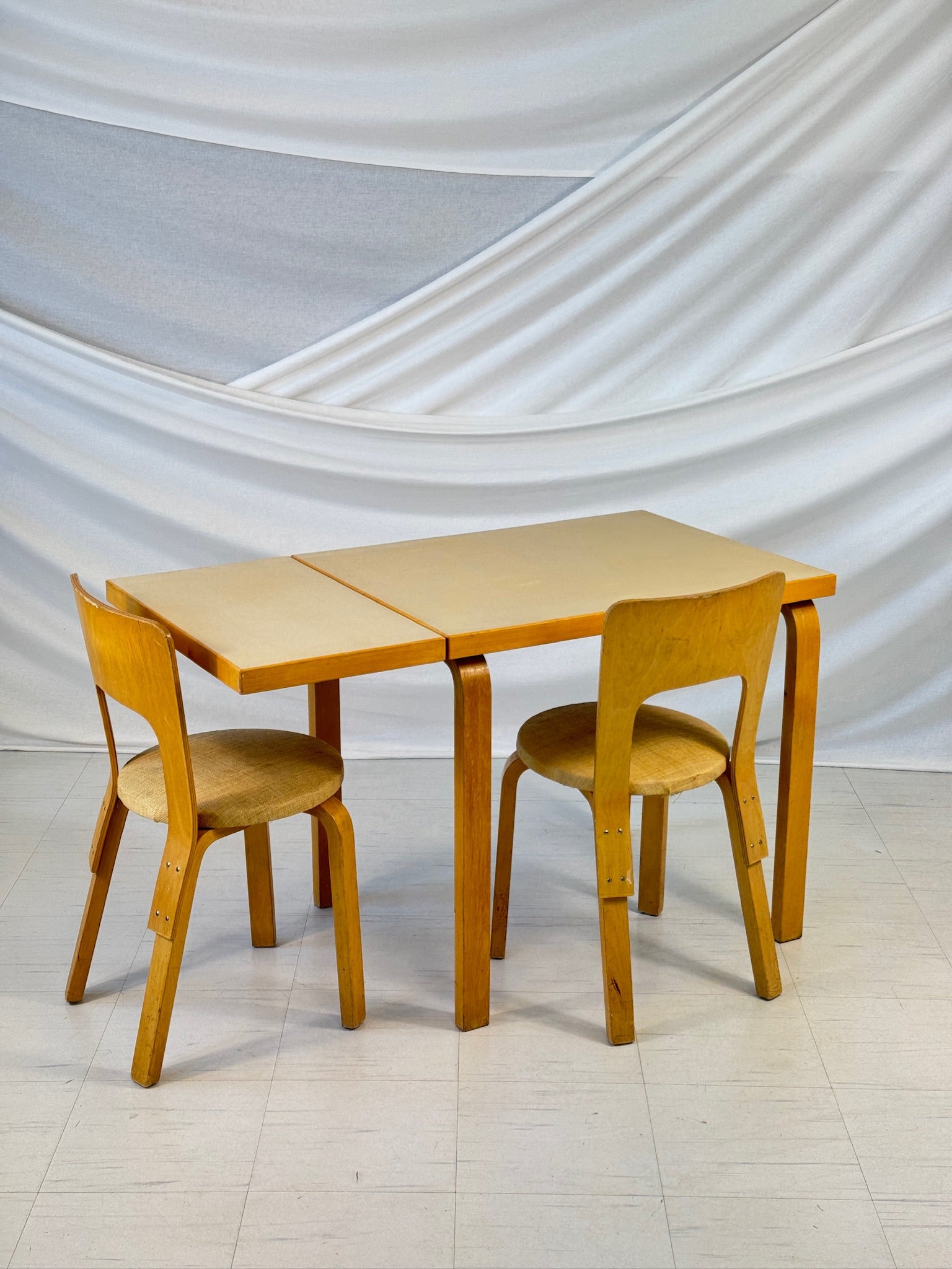 Vintage Artek DL82 table designed by Alvar Aalto, with birch frame and one drop-leaf side extended, photographed against a white draped backdrop.
