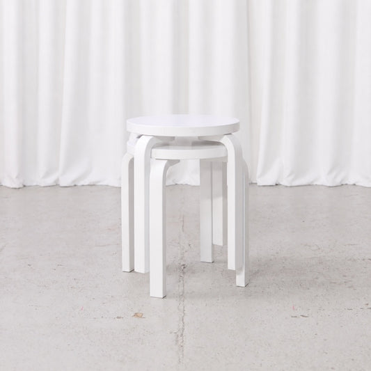 Two white Artek E60 stools by Alvar Aalto stacked in front view